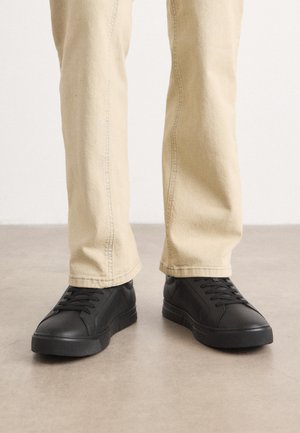 Person trägt beige Hose und schwarze Schnürsneaker und steht auf einem glatten Boden vor einem einfarbigen hellen Hintergrund.