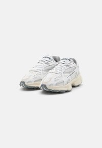 Hvite mesh-sneakers med grå detaljer, teksturerte overflater og en tykk såle. "LACOSTE"-logo vist på tungen og siden.