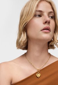 Collar con colgante en forma de corazón dorado y acentos decorativos, con una cadena gruesa y textura lisa, llevado sobre una blusa marrón.