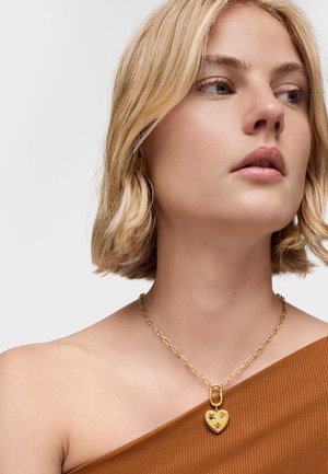 Collar con colgante en forma de corazón dorado y acentos decorativos, con una cadena gruesa y textura lisa, llevado sobre una blusa marrón.