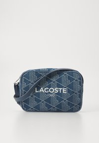 Borsa a tracolla in denim con motivo geometrico, colore blu scuro, hardware argentato e logo bianco "LACOSTE 1933" sulla parte anteriore. Fascia regolabile.