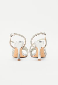 Silberne Sandalen mit Absatz und Knöchelriemen. Riemchen-Design, metallisches Finish und strukturierte Akzente. Offene Rückseite und verstellbarer Schnallenverschluss.