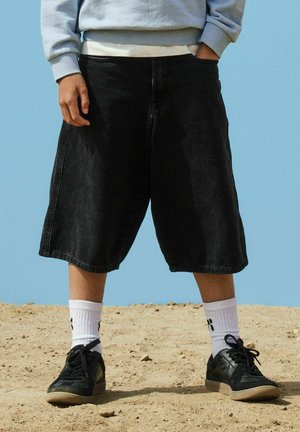 Persoană care stă pe un teren nisipos, purtând pantaloni scurți largi negri, șosete albe cu imprimeu de lăbuțe, adidași negri și un hanorac gri deschis, cu o mână în buzunar.