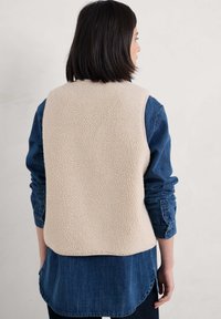 Bež fleecova jakna z mehkim teksturo, oblečena čez modro denim srajco. Jakna nima zapiranja in ima zaobljen rob.