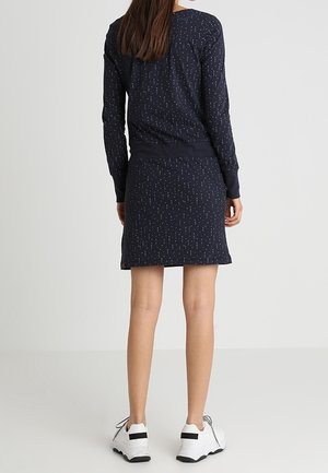 Frau trägt ein marineblaues, langärmeliges Kleid mit kleinen weißen Punkten, kombiniert mit weißen Sneakers, steht vor einem schlichten Hintergrund.
