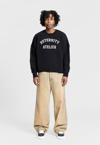 Sort strikket sweatshirt med teksten "RETERNITY ATELIER", parret med beige vide bukser og sorte sneakers med hvide accenter.