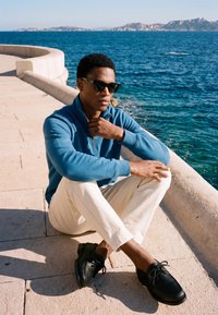 Mann mit Sonnenbrille, blauem Pullover und weißen Hosen, der mit verschränkten Beinen auf Steinpflaster am Meer mit entfernten Bergen sitzt.