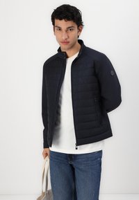Giacca trapuntata blu navy con colletto alto e chiusura con zip, dotata di due tasche laterali e un tessuto testurizzato nella parte superiore.