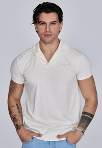 Cremefarvet kortærmet polo-shirt med en knapkrave, der har diskret "SIKSILK" branding og en tætsiddende pasform. Casual denim bukser bæres.