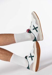 Białe sneakersy z zielonymi akcentami, beżowym zamszem i teksturowaną podeszwą. Sznurowany fason, z czerwoną łatą z logo i prążkowanymi białymi skarpetkami.