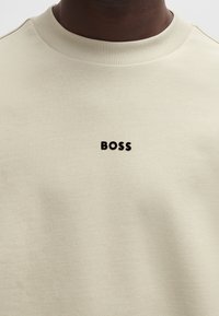 Beige sweatshirt tillverkad av mjukt tyg, med en ribbad crew neck och en svart "BOSS"-logotyp placerad på bröstet.