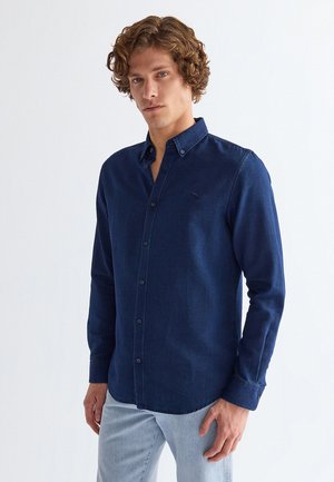 Junger Mann mit lockigem Haar, der ein dunkelblaues, geknöpftes Langarmhemd und hellblaue Jeans vor einem schlichten Hintergrund trägt.