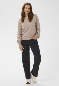 Femme portant un pullover beige, un jean noir et des baskets blanches, se tenant avec une main dans la poche devant un fond blanc uni.