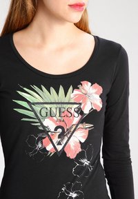 Schwarzes Langarmshirt mit floralem Design in Rosa- und Grüntönen, zentriertes dreieckiges Logo mit dem Schriftzug "GUESS" und Verzierungen.