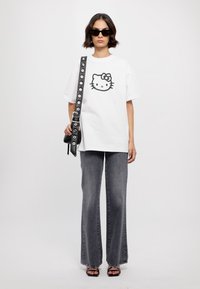 Tricou alb cu grafică neagră Hello Kitty, asortat cu blugi gri evazați și o geantă neagră cu gromete argintii. Ochelari de soare negri purtați.
