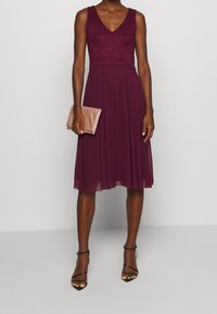 Burgundské šaty s krajkovým korzetem, V-výstřihem a plisé sukní, doplněné o světle růžovou kabelku a černé sandály na podpatku.