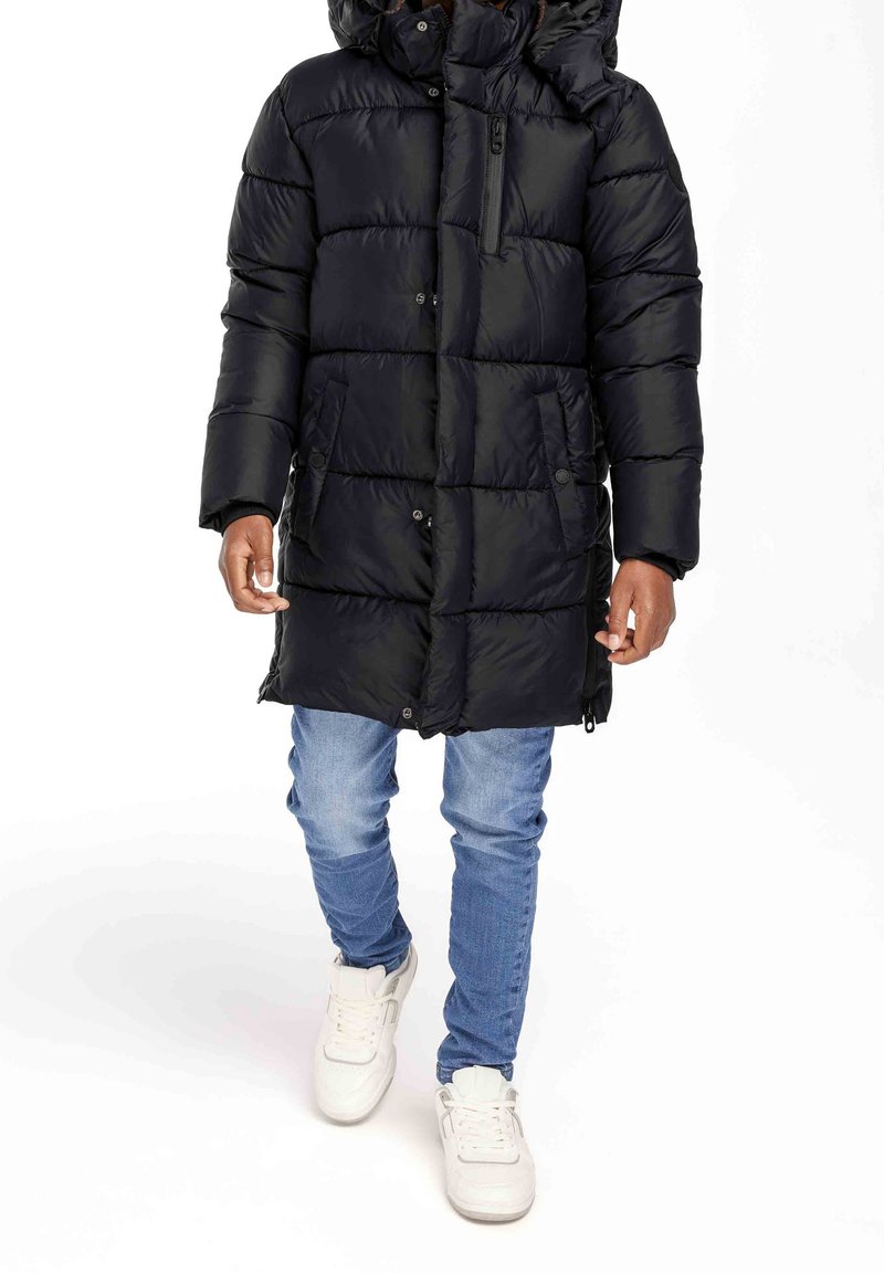 Personne portant un long manteau d'hiver noir matelassé avec des poches latérales, un jean bleu et des baskets blanches, debout sur un fond blanc uni.