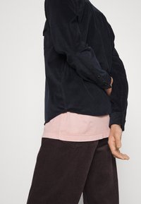 Granatowa sztruksowa koszula z mankietami na guziki, nałożona na jasnoróżową koszulę, w połączeniu z ciemnobrązowymi spodenkami z prążkowanej tkaniny, podkreślającymi fakturę i dopasowanie.