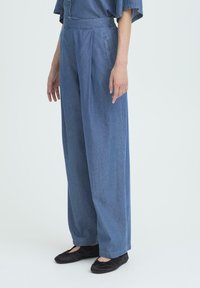Vrouw in losse, hooggemiddelde blauwe denimbroek met plooien en zijzakken, gecombineerd met zwarte balletschoenen, staand tegen een witte achtergrond.
