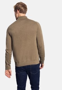 Maglione marrone a maglia con colletto alto e orlo a coste, indossato sopra jeans blu scuri. Il tessuto ha una texture liscia.