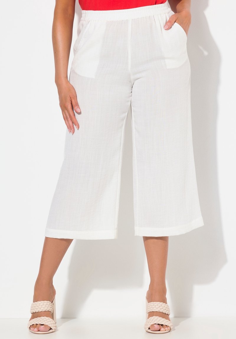 Femme portant un pantalon blanc ample et court avec taille élastique et poches latérales, associé à des sandales beige à brides tressées.