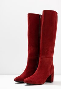 Czerwone zamszowe kozaki za kolano na masywnej szpilce i z zamkiem błyskawicznym z boku. Gładka tekstura, spiczasty nosek i dopasowany krój tworzą elegancki wygląd.
