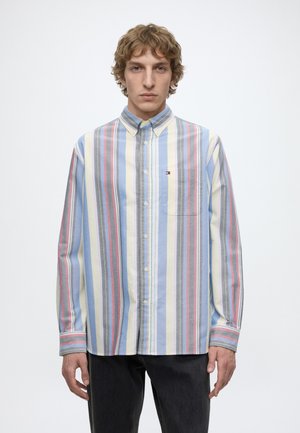 Jeune homme aux cheveux bouclés portant une chemise à manches longues boutonnée avec des rayures verticales bleues, grises, jaunes et rouges, debout devant un fond uni.