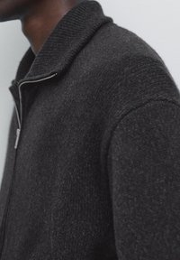 Black zip-up sweater with a ribbed collar and textured fabric, featuring a smooth front and slightly raised knit shoulders. Casual design.