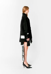 Chaqueta negra de gran tamaño con bolsillos, acentos blancos en contraste, diseño en capas, llevada sobre una falda corta negra. Zapatos negros de punta con herrajes decorativos.