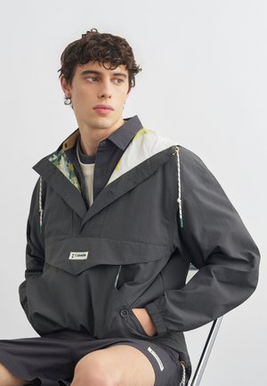 Joven con cabello rizado que lleva una chaqueta Columbia gris oscuro con capucha floral, sentado y mirando hacia un lado contra un fondo liso.