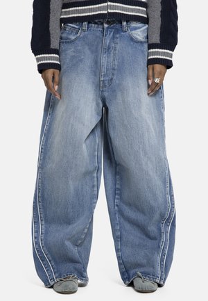 Person trägt weite, ausgebleichte blaue Jeans mit weißen Seitenstreifen, kombiniert mit einem marineblau-grau gestreiften Pullover und grauen Schuhen.