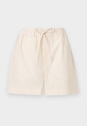 Femme debout portant une chemise rose clair à manches longues, un short beige à cordon de serrage et des sandales beiges avec détails en nœud, sur un fond uni.