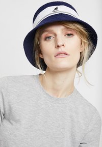 Chapeau seau en éponge bleue avec des accents de rayures blanches et un petit logo. Texture douce et forme arrondie, porté avec un T-shirt gris.