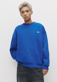 Sweat-shirt bleu avec un col rond et des épaules tombantes, arborant un petit logo blanc sur la poitrine et des poignets côtelés. Tissu doux.