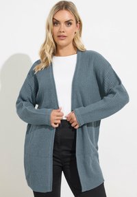 Cardigan tricoté bleu clair avec une texture côtelée, à l'avant ouvert et à manches longues. Associé à un haut blanc et un pantalon noir, avec une coupe décontractée.