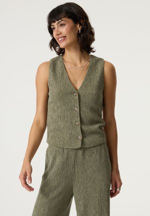 Mujer sonriendo, con un chaleco sin mangas de botón texturizado en verde oliva y pantalones de pierna ancha a juego, accesorios con collares en capas y anillos.