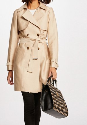 Vrouw draagt een beige trenchcoat met riem en zwarte broek, houdt een grote zwart-beige handtas met houndstooth-patroon vast.