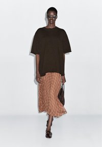 Modelka nosi czarny, oversize'owy top z krótkim rękawem, brązową, w kratę midi spódnicę, czarne okulary przeciwsłoneczne, trzyma w ręku małą czarną torebkę i idzie do przodu.