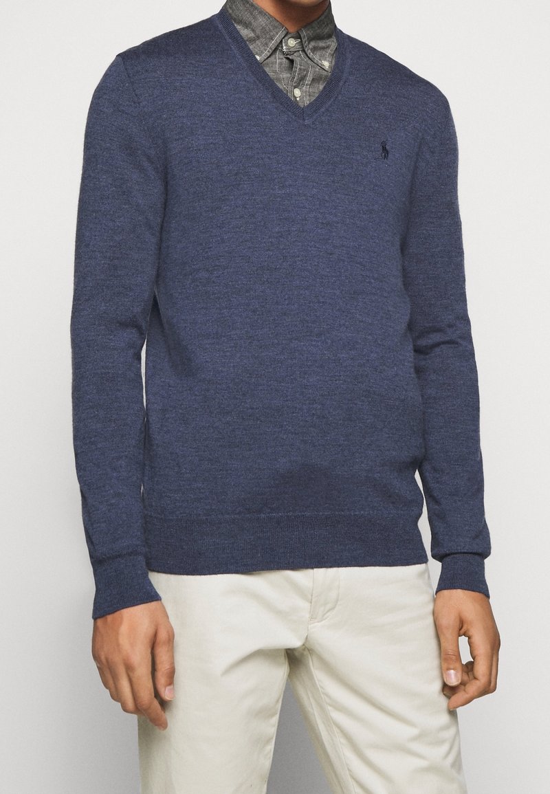 Pull en maille texturée bleu marine à col en V, avec un petit logo sur la poitrine, associé à une chemise grise à boutons en dessous.