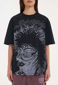 Černé oversized tričko s velkým šedým potiskem lebky s dlouhými vlasy a řetězovými akcenty, doplněné stylizovaným textem nahoře.