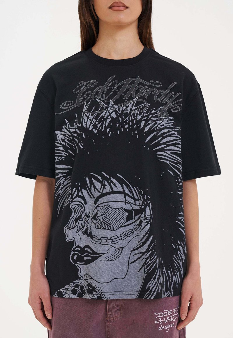 Černé oversized tričko s velkým šedým potiskem lebky s dlouhými vlasy a řetězovými akcenty, doplněné stylizovaným textem nahoře.