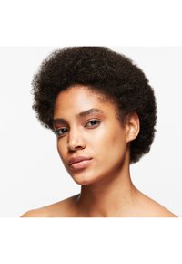Gros plan sur une personne avec des cheveux courts et bouclés, une peau lisse, un maquillage naturel et un teint uniforme sur fond blanc.