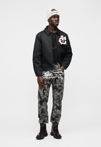 Homme portant une veste noire avec un motif blanc, un pantalon gris à motifs avec un design blanc, des bottes marron et un bonnet blanc, debout devant un fond uni.