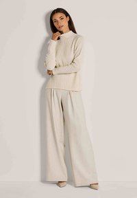 Crème ribget turtleneck sweater gecombineerd met losse, lichtbeige wijde broek en neutraalgekleurde hakken, tegen een neutrale achtergrond.