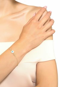 Gold chain bracelet with an eye-shaped charm featuring a blue center, small clear stones, and a delicate link design. Smooth skin tone and soft fabric.