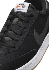 Musta mokkanahkainen tennari, jossa on valkoinen kumipohja ja mustat nahkaiset yksityiskohdat. Varustettu tasaisilla mustilla nauhoilla ja valkoisella kielenauhalla, jossa oranssi logo.