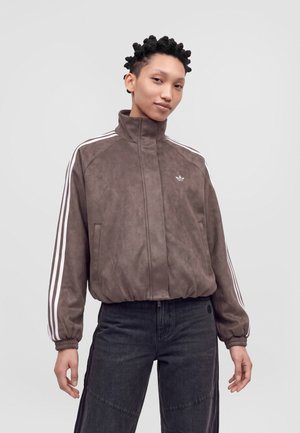 Modèle portant une veste Adidas marron à fermeture éclair, ornée de bandes blanches sur les manches, et un jean foncé, devant un fond de studio gris clair.
