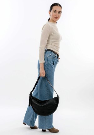 Black leather handbag with a curved shape, held by a person wearing a light beige fitted top and wide blue jeans. Smooth texture, minimal hardware.