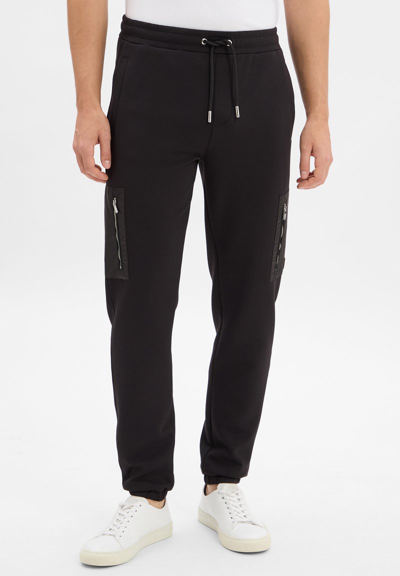 Zwarte joggerbroeken met elastische tailleband, ritszakken aan de zijkanten, ribgebreide boorden, gemaakt van zachte stof, gecombineerd met witte sneakers.