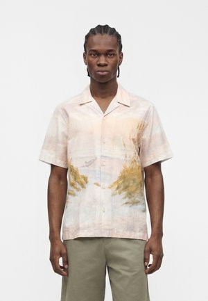 Man with braided hair wears short-sleeve button-up shirt with soft watercolor landscape print and light olive green shorts against plain background.